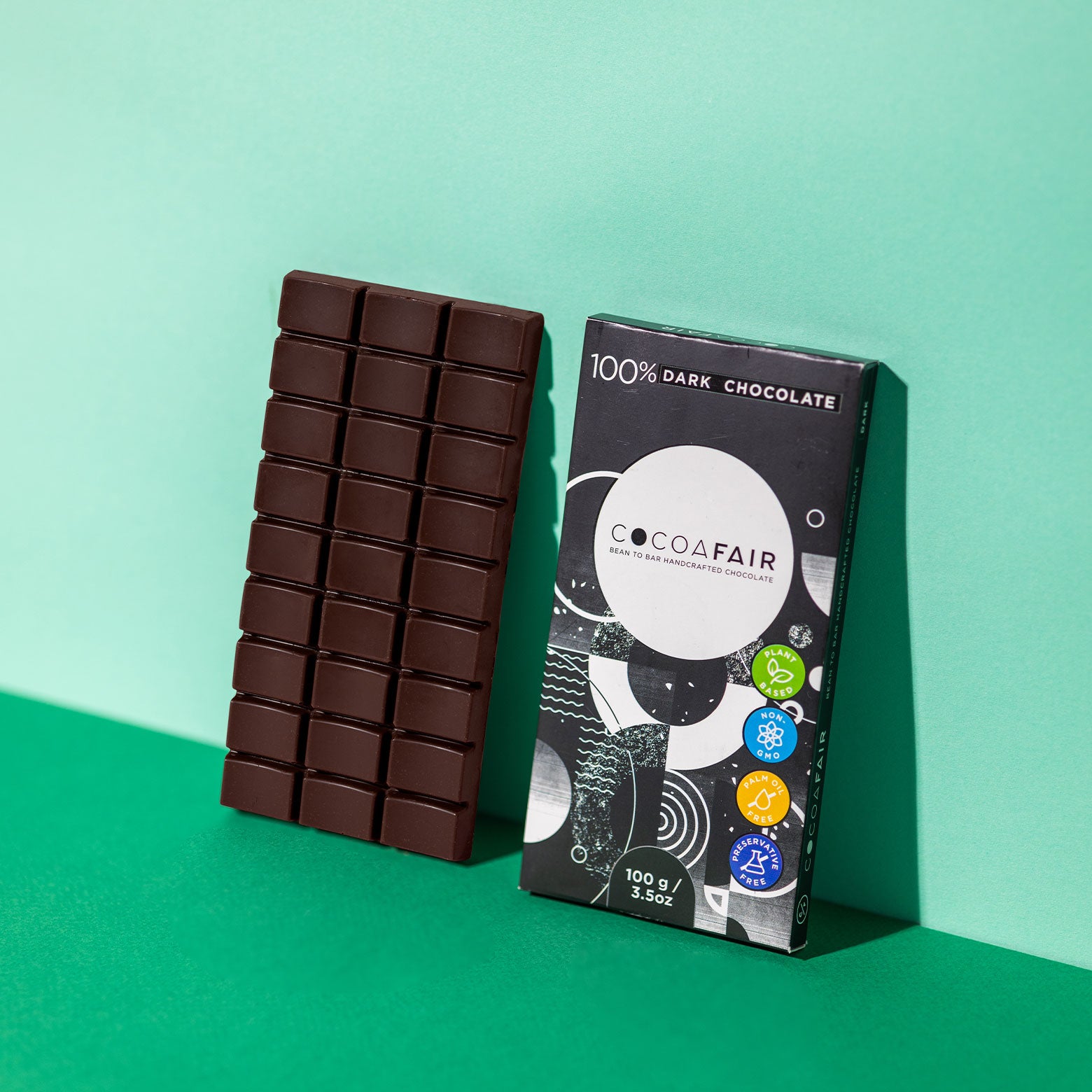 COCOAFAIR 100g 100% Dark Chocolate. Lifestyle image. Black packaging on green background. Chocolate slab leans against green background and black packaging leans against green background at a angle.