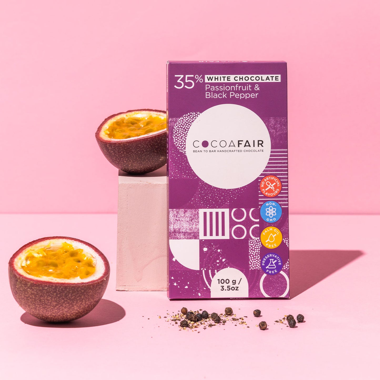 COCOAFAIR 100g 35% White Chocolate Lifestyle shot - Purple packaging on a pink  background with passionfruit in halve with black pepper corns. Passionfruit & Black Pepper flavoured chocolate. 