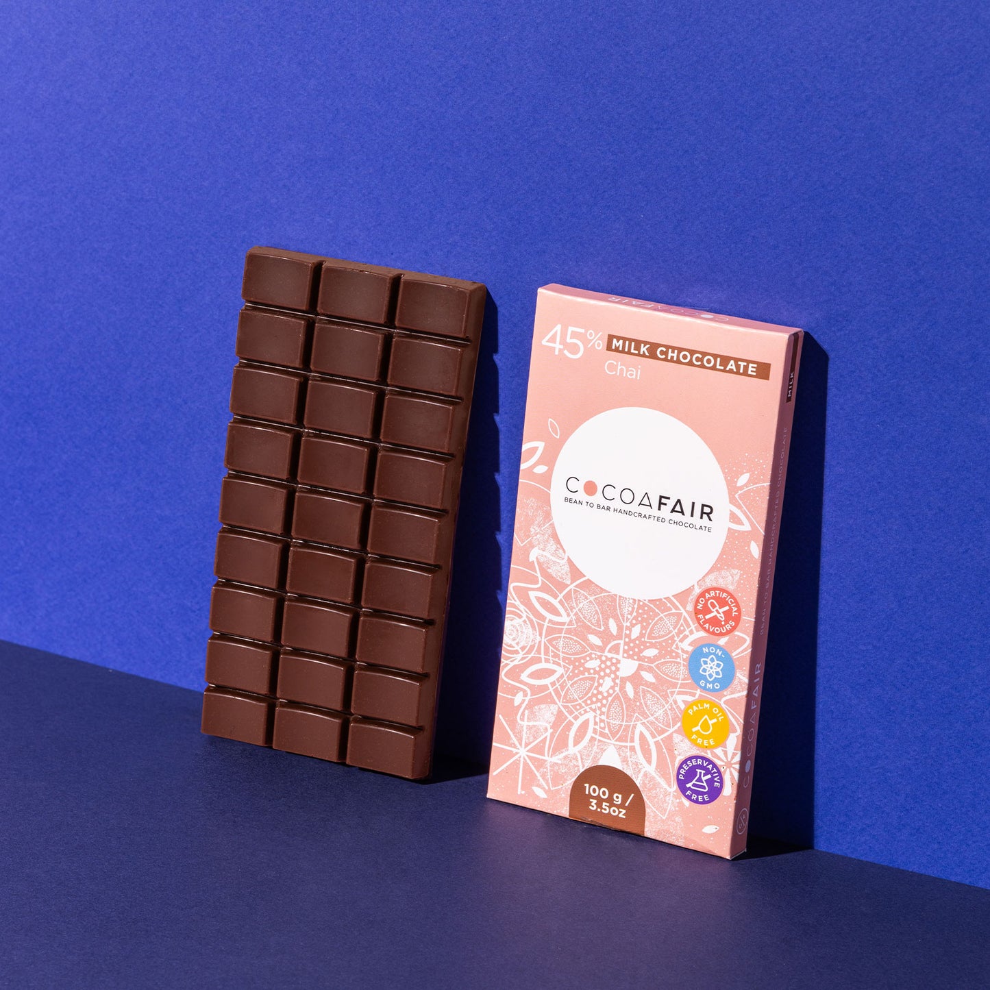 COCOAFAIR 100g 45% Milk Chocolate Lifestyle shot - Pink packaging on navy blue background with a slab of milk chocolate. Chai flavoured chocolate