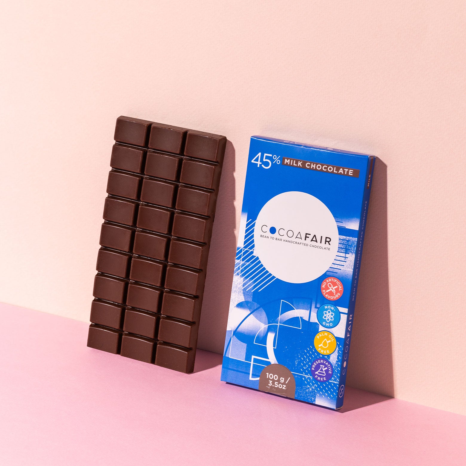 COCOAFAIR 100g 45% Milk Chocolate Lifestyle shot - Blue packaging with milk chocolate slab at a angle on pink background.