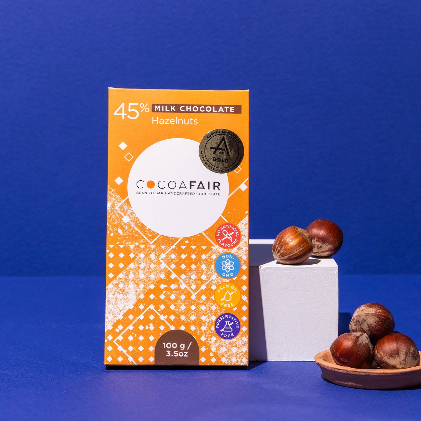 COCOAFAIR 100g 45% Milk Chocolate Lifestyle shot - Orange packaging with Hazelnuts around on navy blue  background. Hazelnut flavoured chocolate