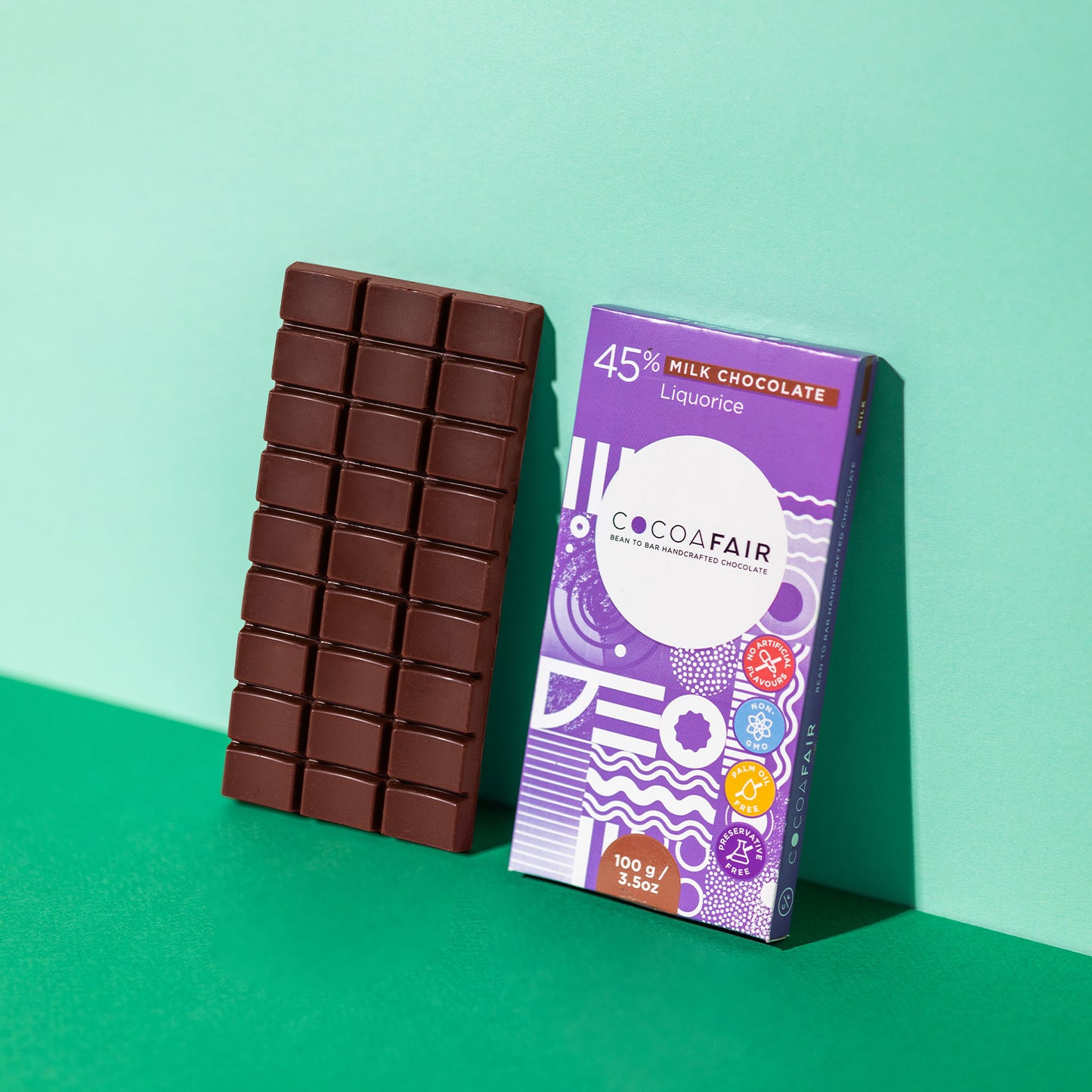 COCOAFAIR 100g 45% Milk Chocolate Lifestyle shot - Purple packaging on green background with a slab of milk chocolate. Liquorice flavoured chocolate