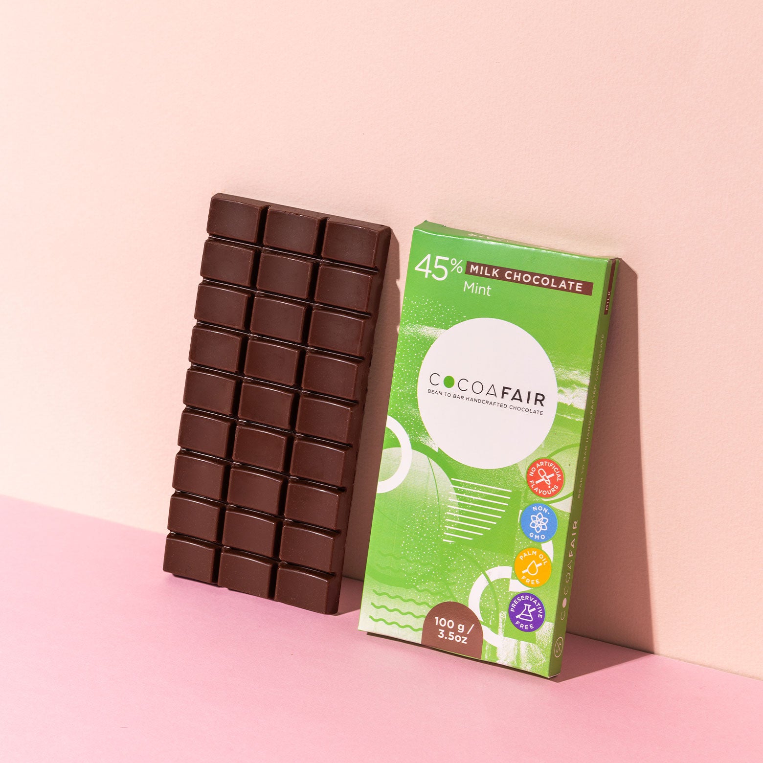 COCOAFAIR 100g 45% Milk Chocolate Lifestyle shot - Green packaging  with milk chocolate slab on pink background at a angle. Mint flavoured chocolate.