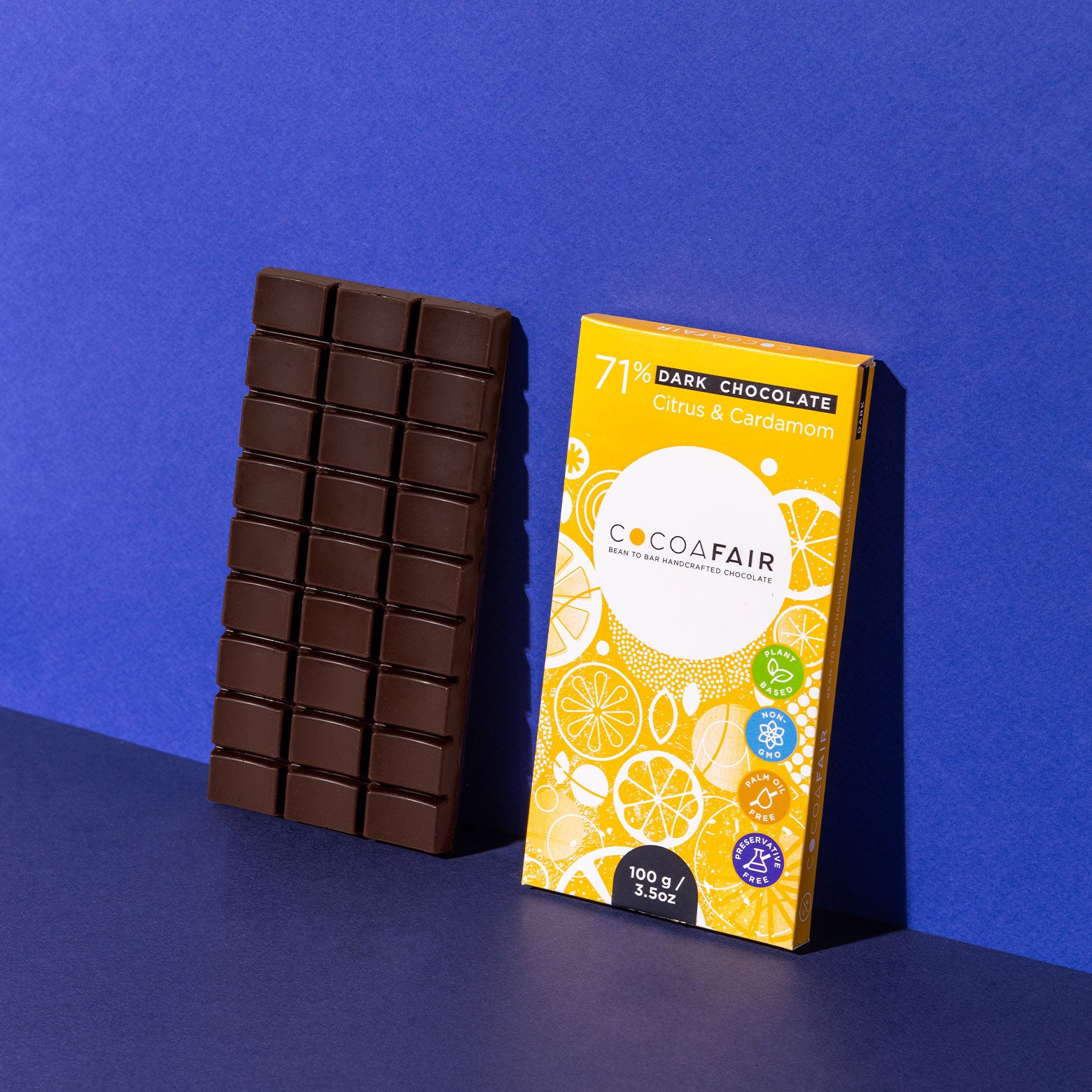 COCOAFAIR 100g 71% Dark Chocolate LIfestyle shot - Yellow packaging with dark chocolate slab at a angle on navy blue background. Citrus and Cardamom flavoured chocolate. 