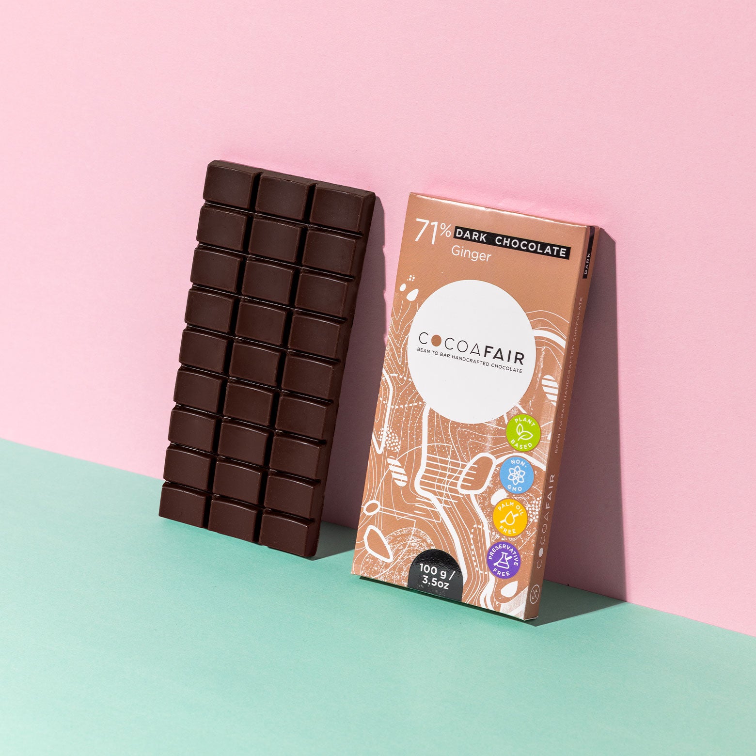 COCOAFAIR 100g 71% Dark Chocolate Lifestyle shot - Brown packaging with dark chocolate slab at a angle on pink and green background. Ginger flavoured chocolate