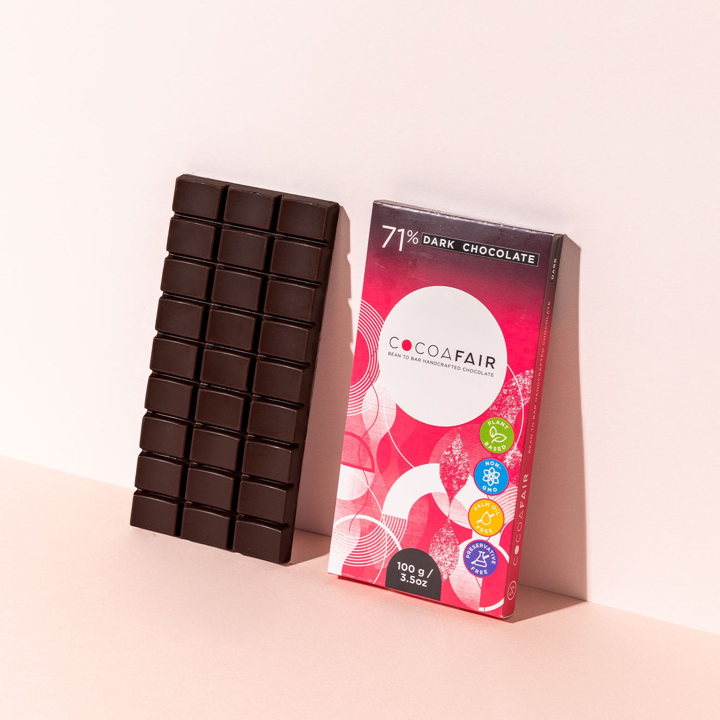 COCOAFAIR 100g 71% Dark Chocolate Lifestyle shot - Pink packaging with dark chocolate slab at a angle on pink background. 