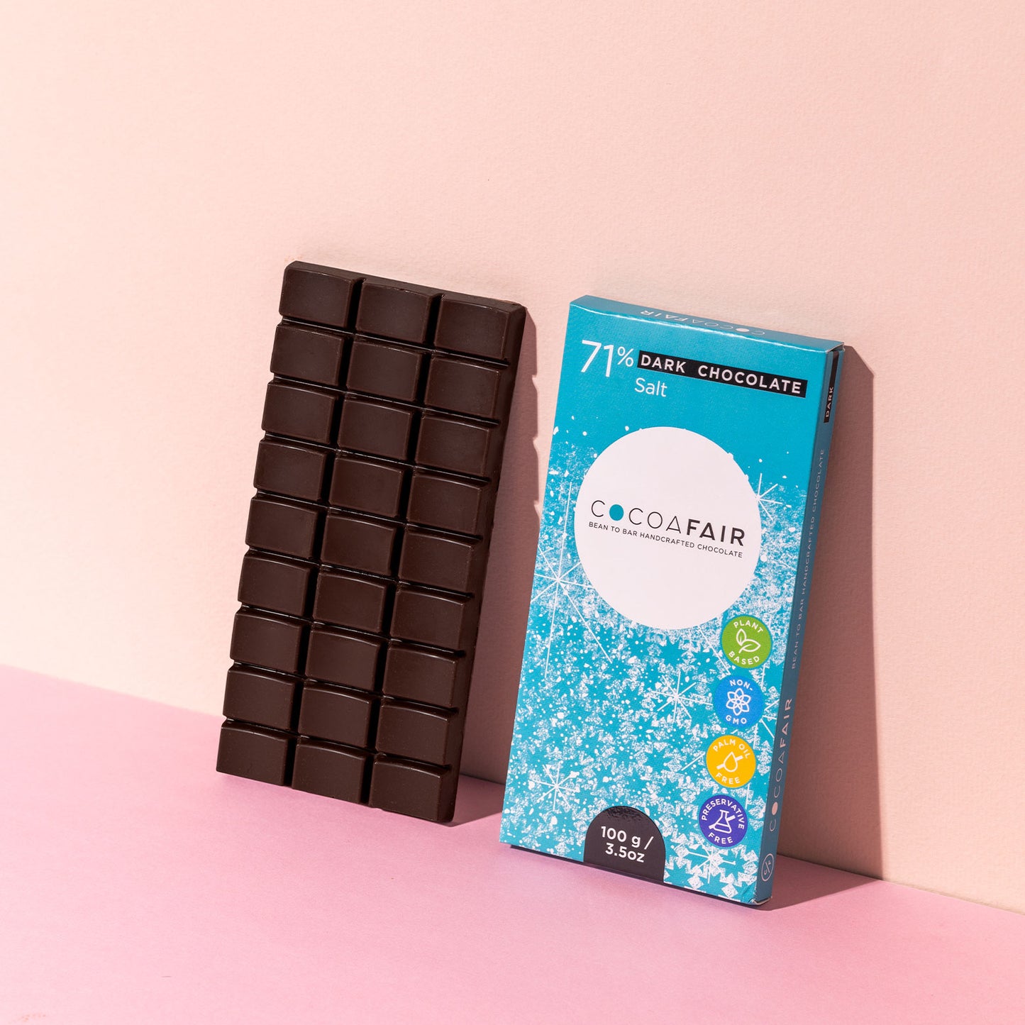 COCOAFAIR 100g 71% Dark Chocolate Lifestyle shot - Blue packaging on pink background with a slab of dark chocolate. Salt flavoured chocolate